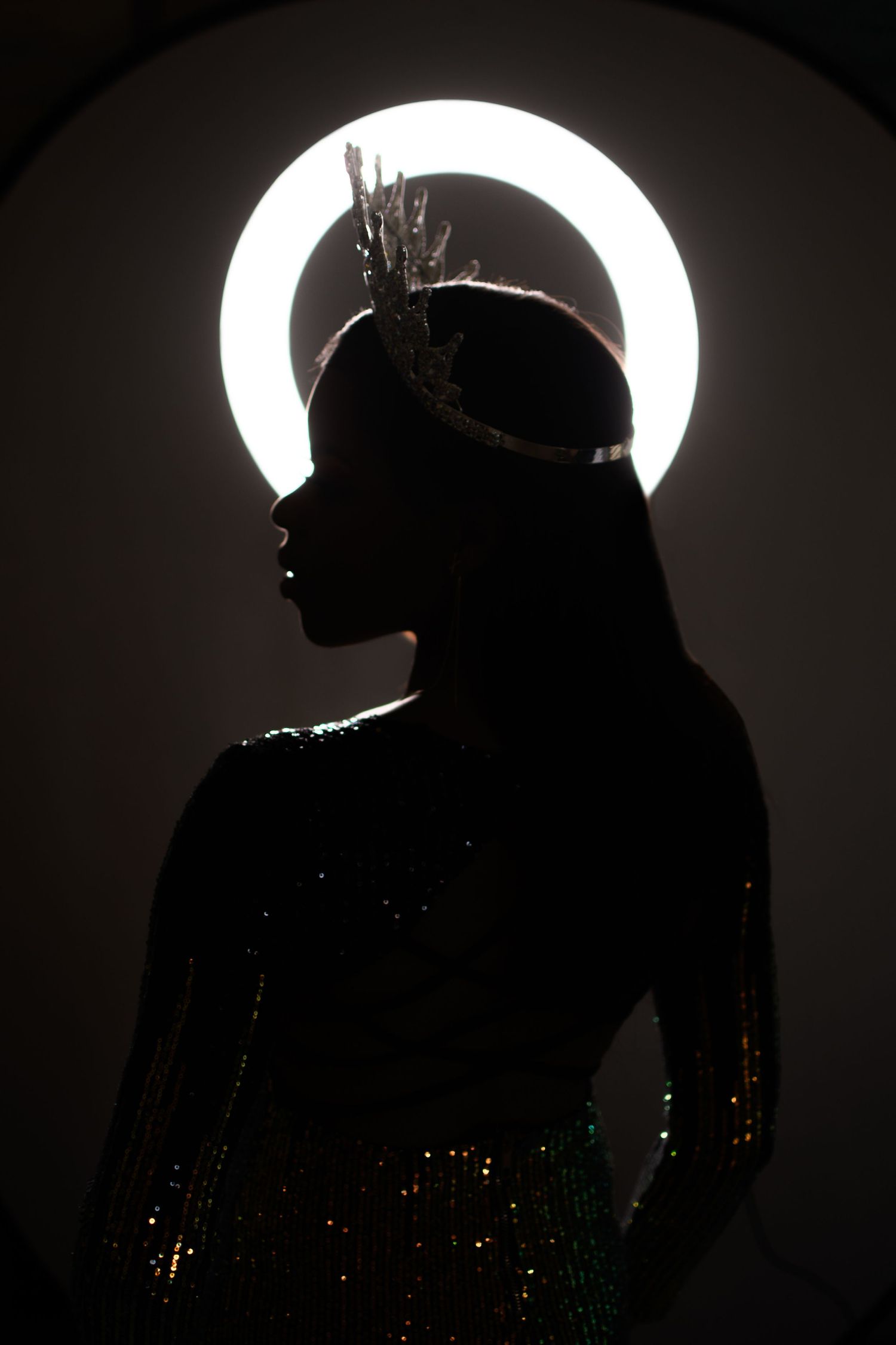 Silhouette of crowned woman in sequin dress posed before glowing circular halo light in dark studio.