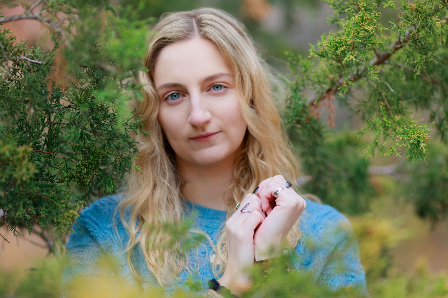 Young blonde woman with blue eyes holds small object while standing among lush green bushes outdoors.