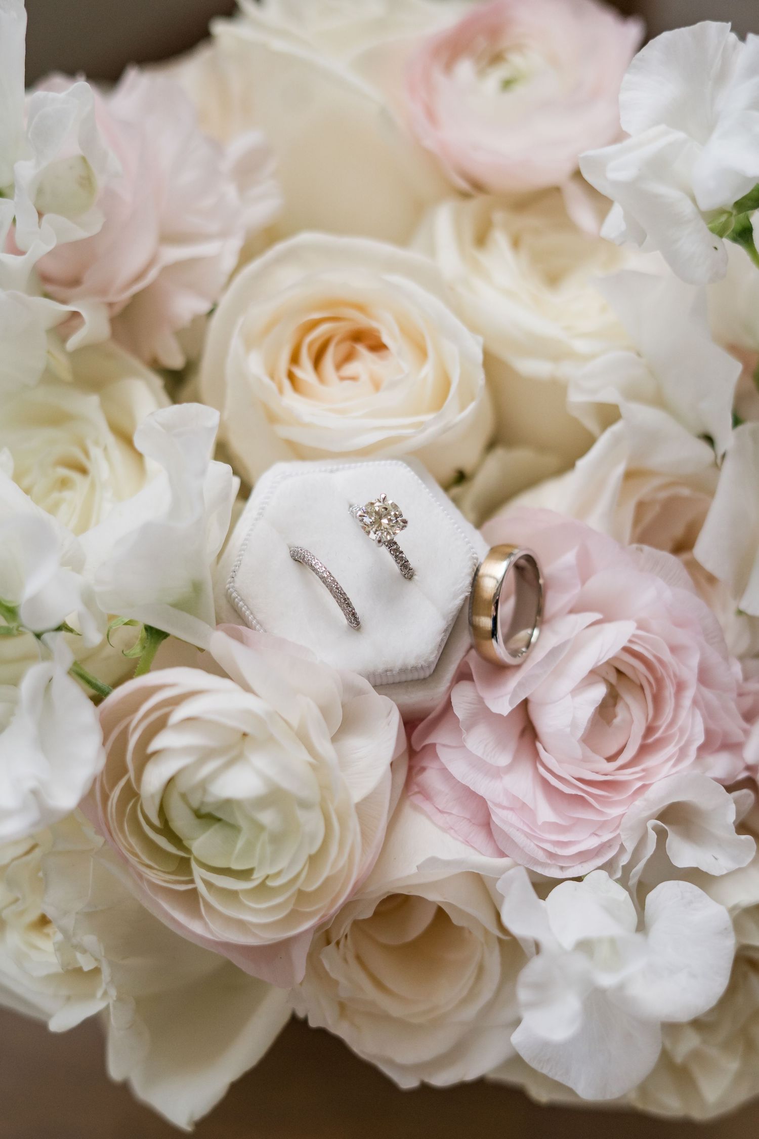Wedding rings displayed in a velvet hexagon box nestled among white and blush ranunculus and roses bouquet.