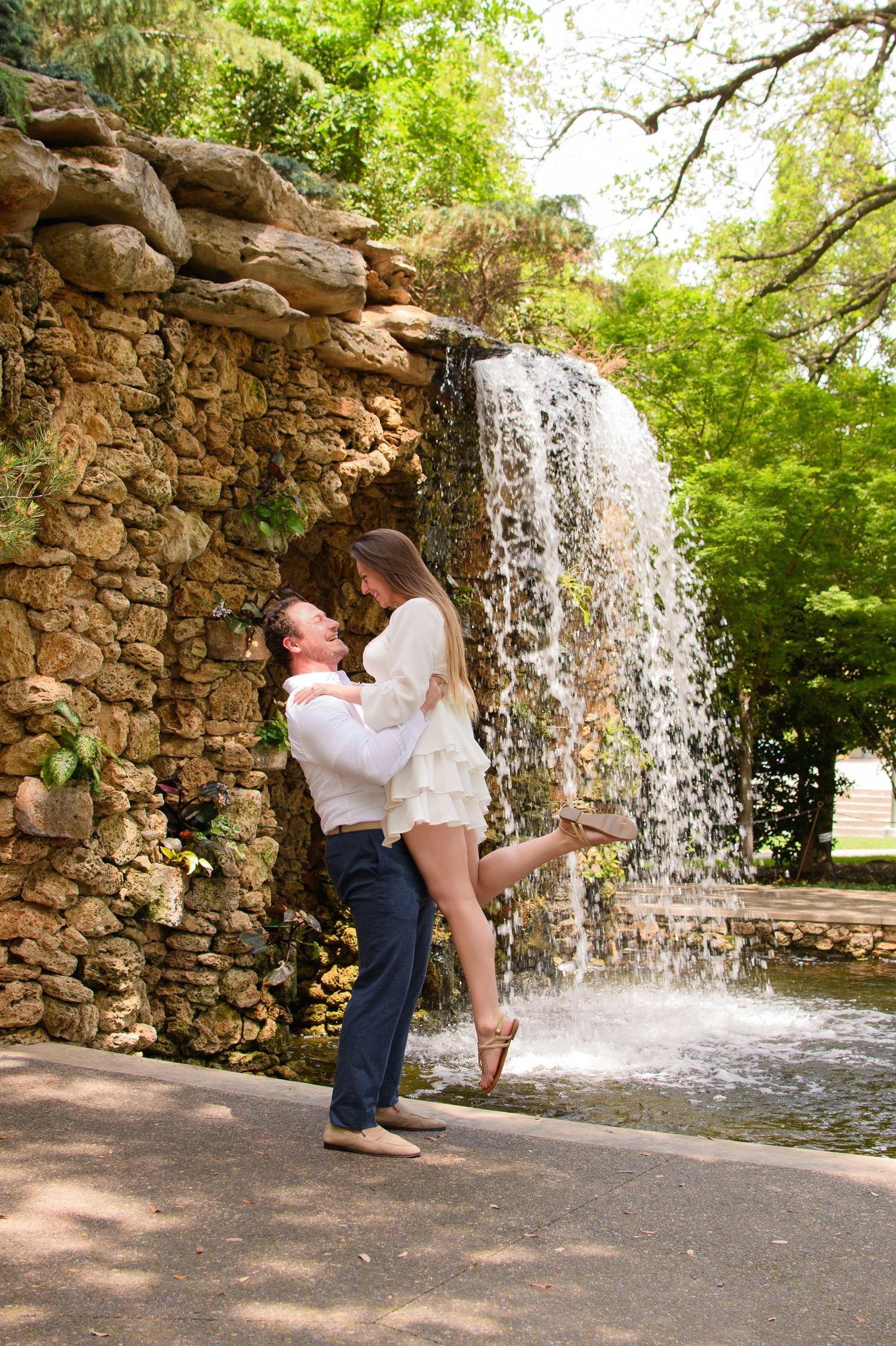 Smiling couple embracing as a man lifts a woman in a white dress beside a stone waterfall in a lush green garden.