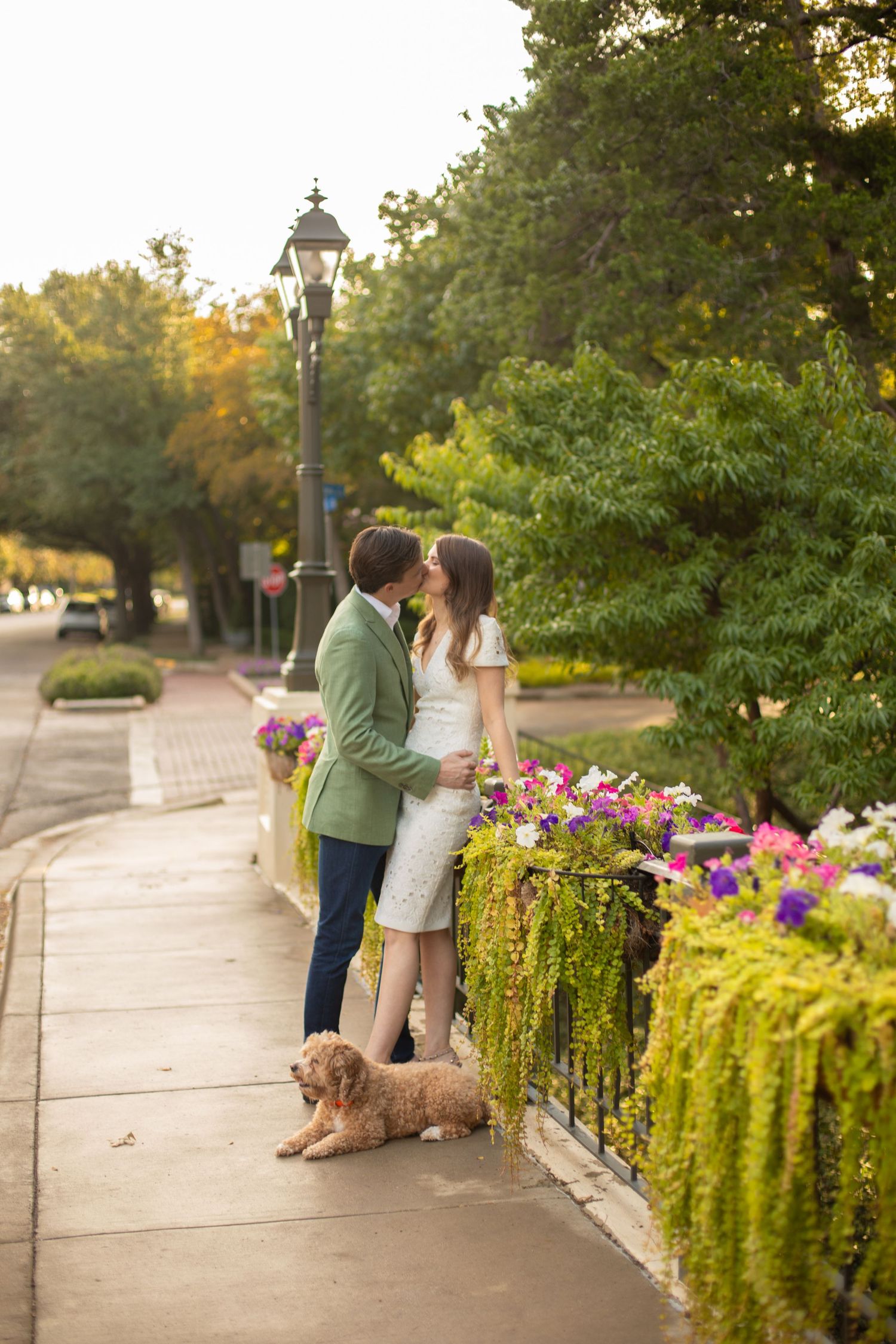 Couple sharing romantic moment beside vibrant flower display with dog on sidewalk.