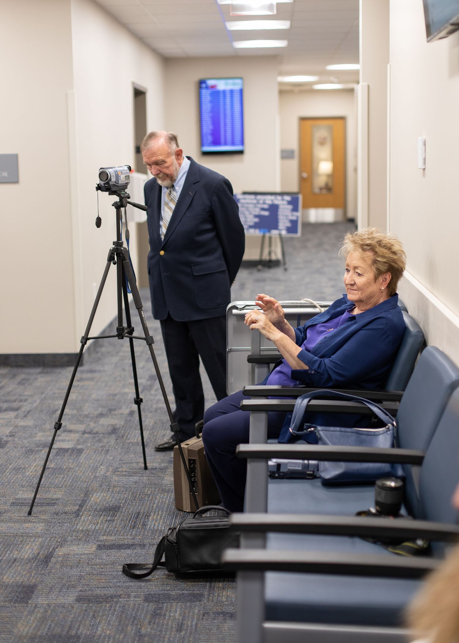 Professional camera setup on a tripod in an office hallway with people waiting in chairs.