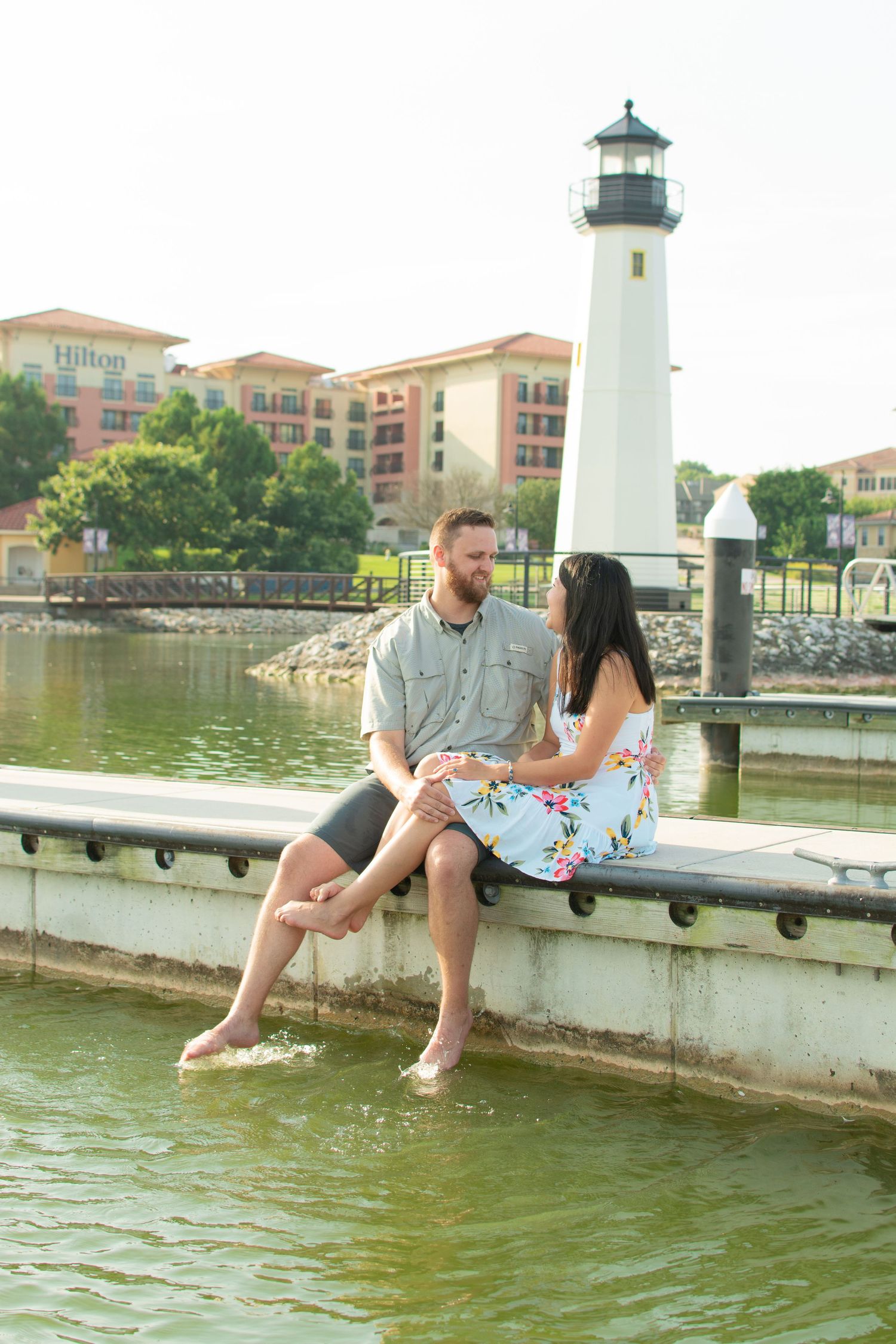 Couple sitting on a waterfront dock with feet in water, lighthouse and Hilton hotel in background.