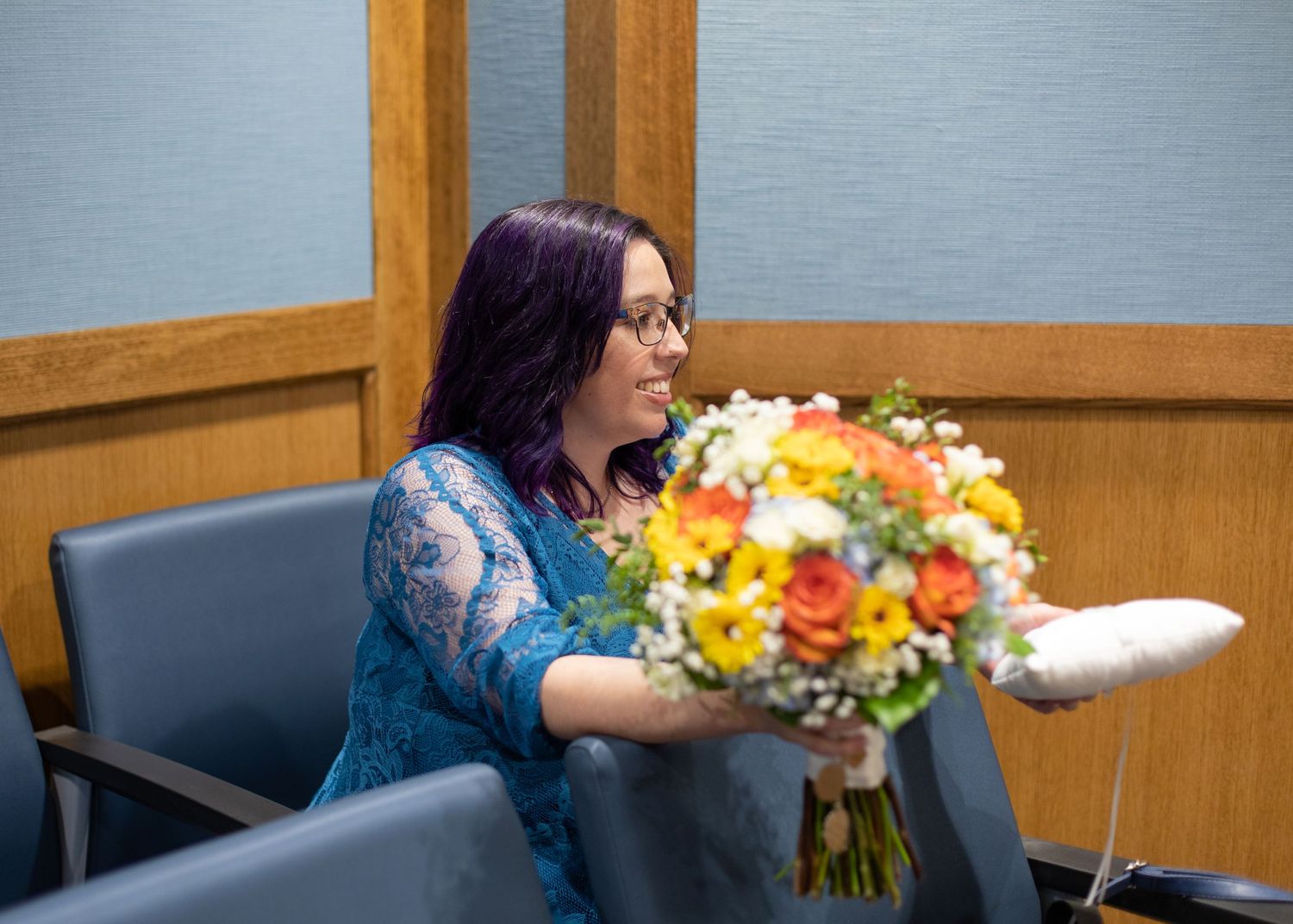 A person in a blue lace dress holds a vibrant bouquet of colorful flowers while seated in a chair.