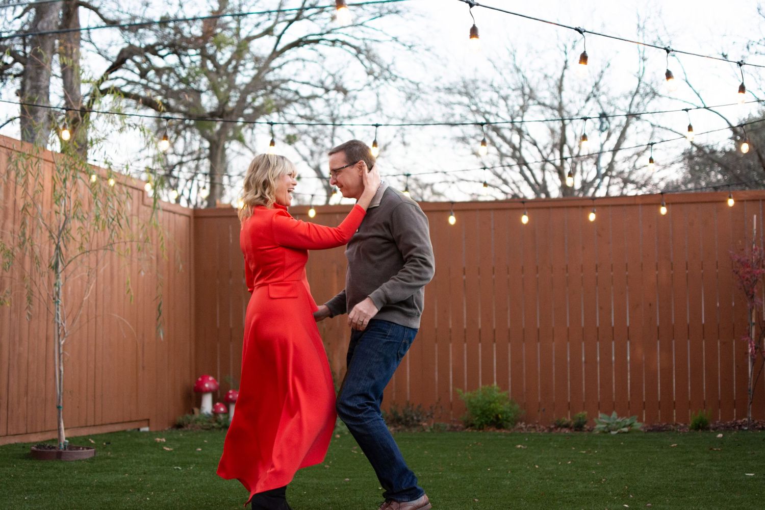 Couple dancing outdoors in backyard with string lights and wooden fence.