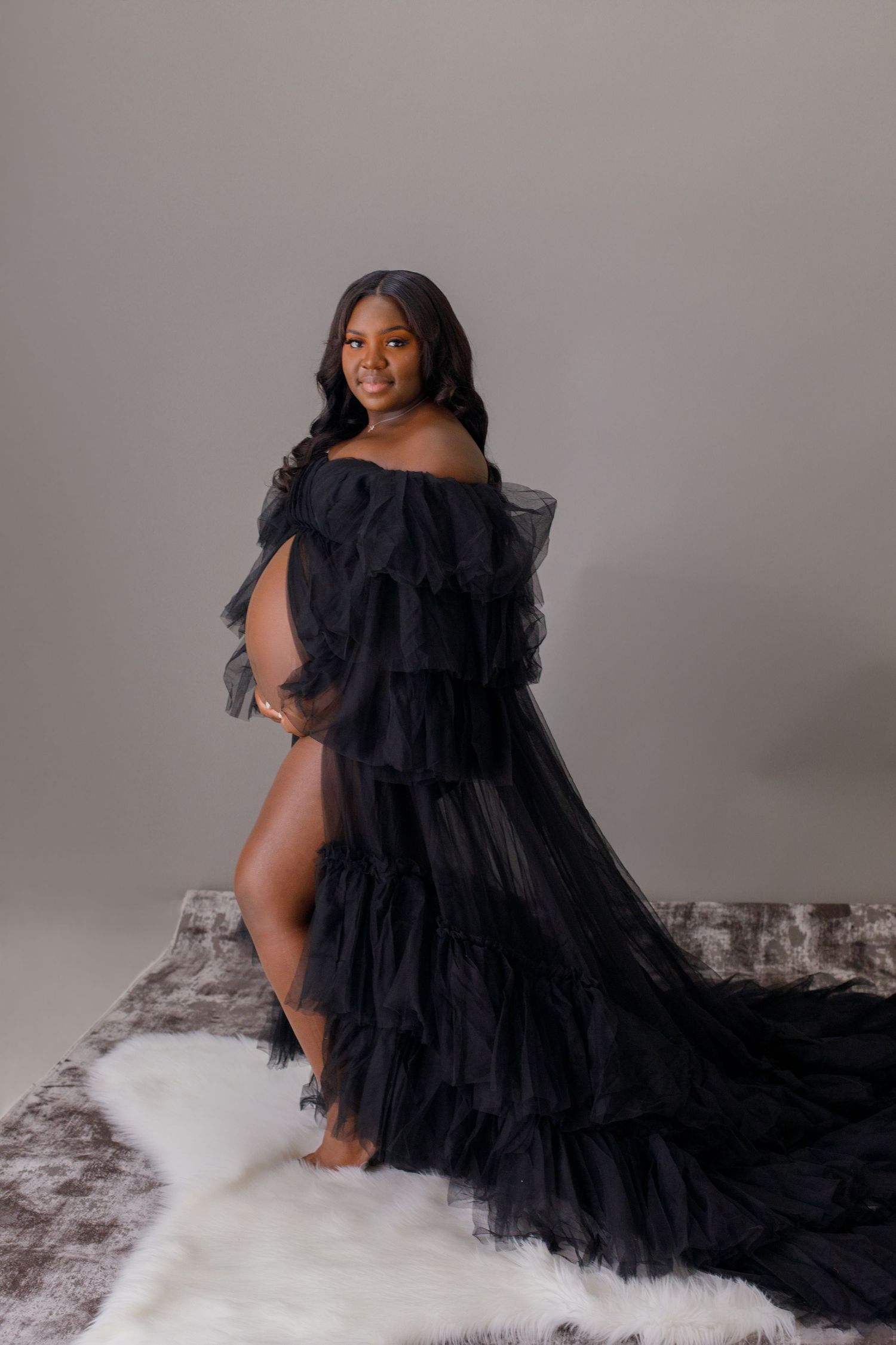 Pregnant Black woman poses elegantly in flowing black ruffled tulle maternity gown on fur rug.