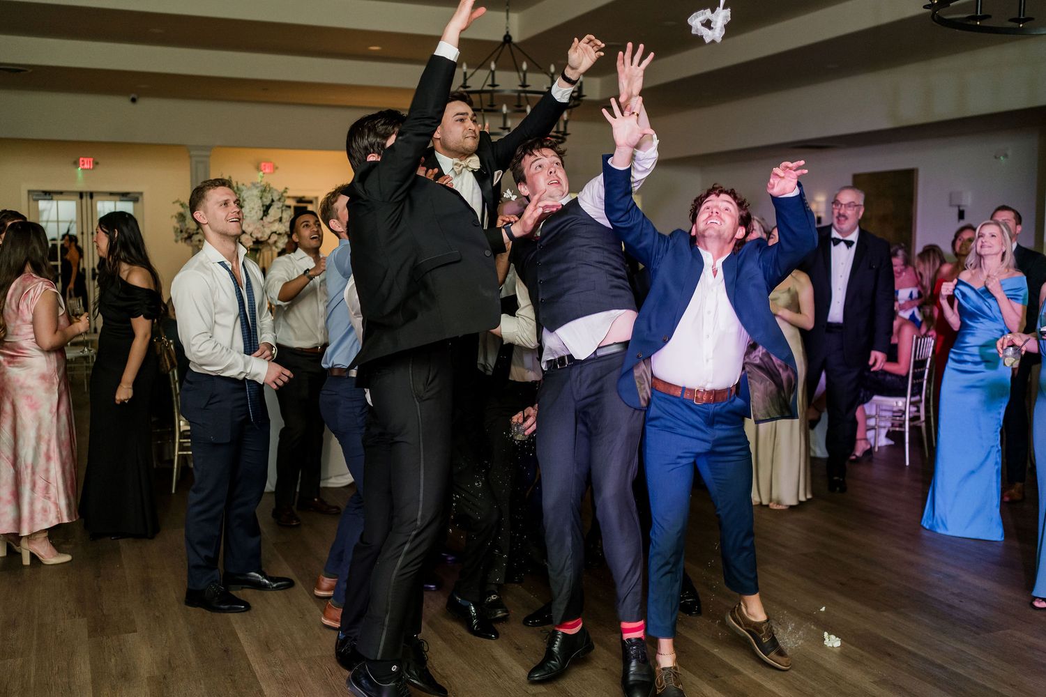 Men in suits jumping and catching bouquet at a lively wedding reception dance floor celebration.
