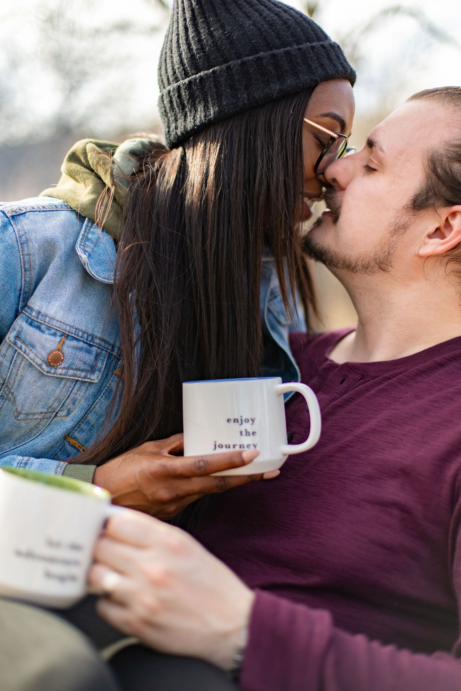 Interracial couple kissing outdoors while holding camping mugs reading enjoy the journey.