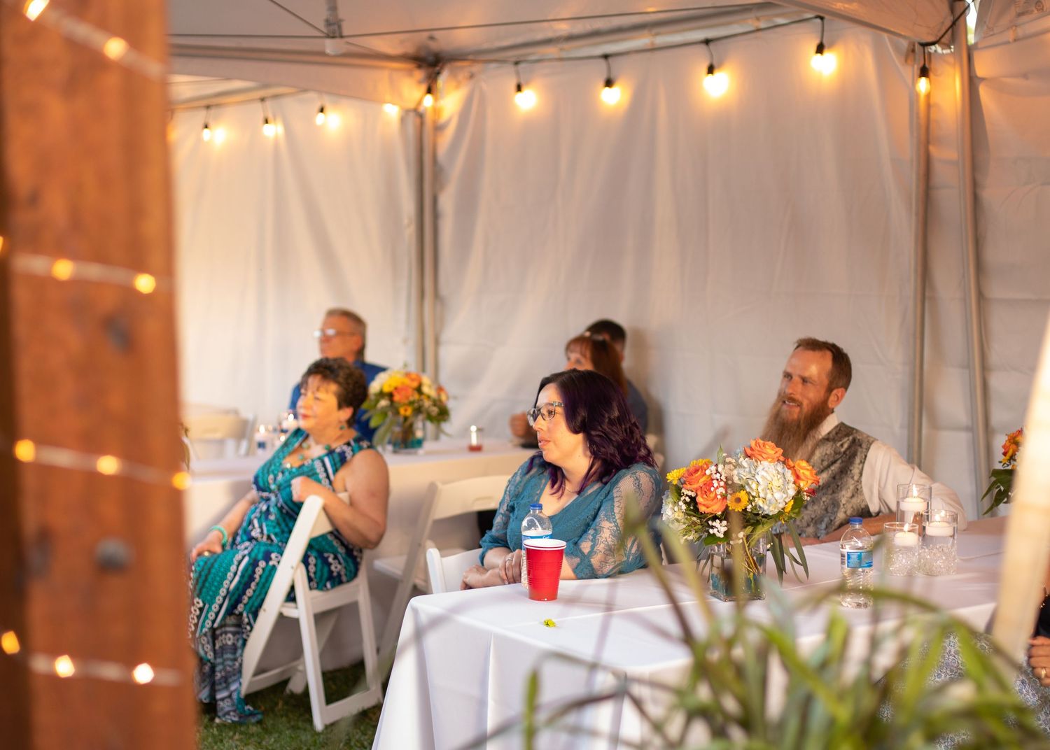 Wedding reception guests dining under string lights in a tented area.