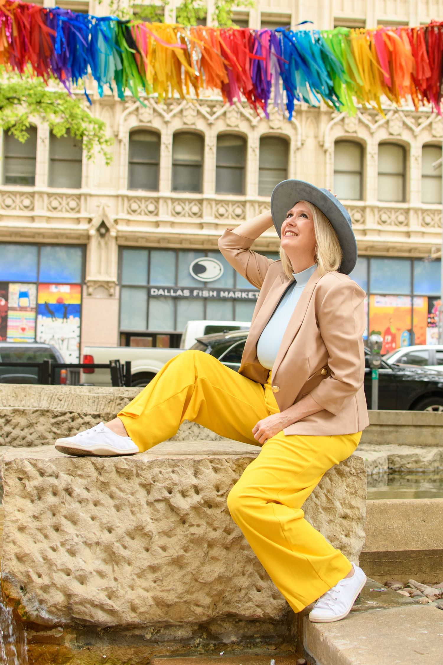 Stylish blonde woman in yellow pants and tan blazer sits by fountain near Dallas Fish Market.