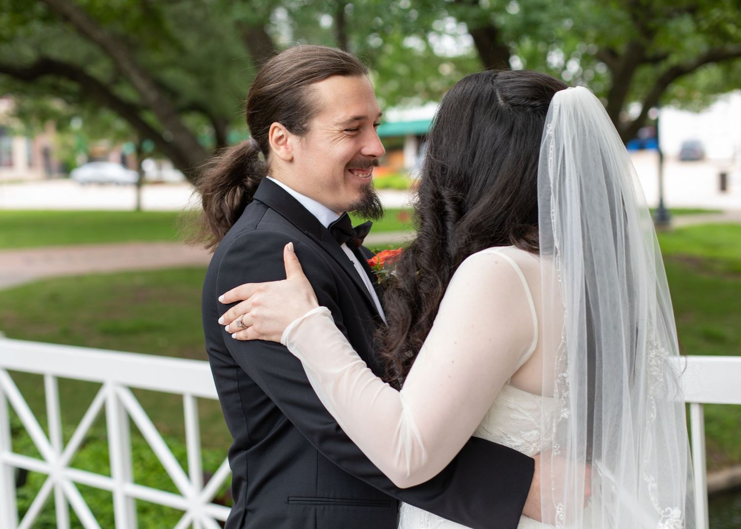 A couple poses together on a red and white bridge over water in a lush garden setting.