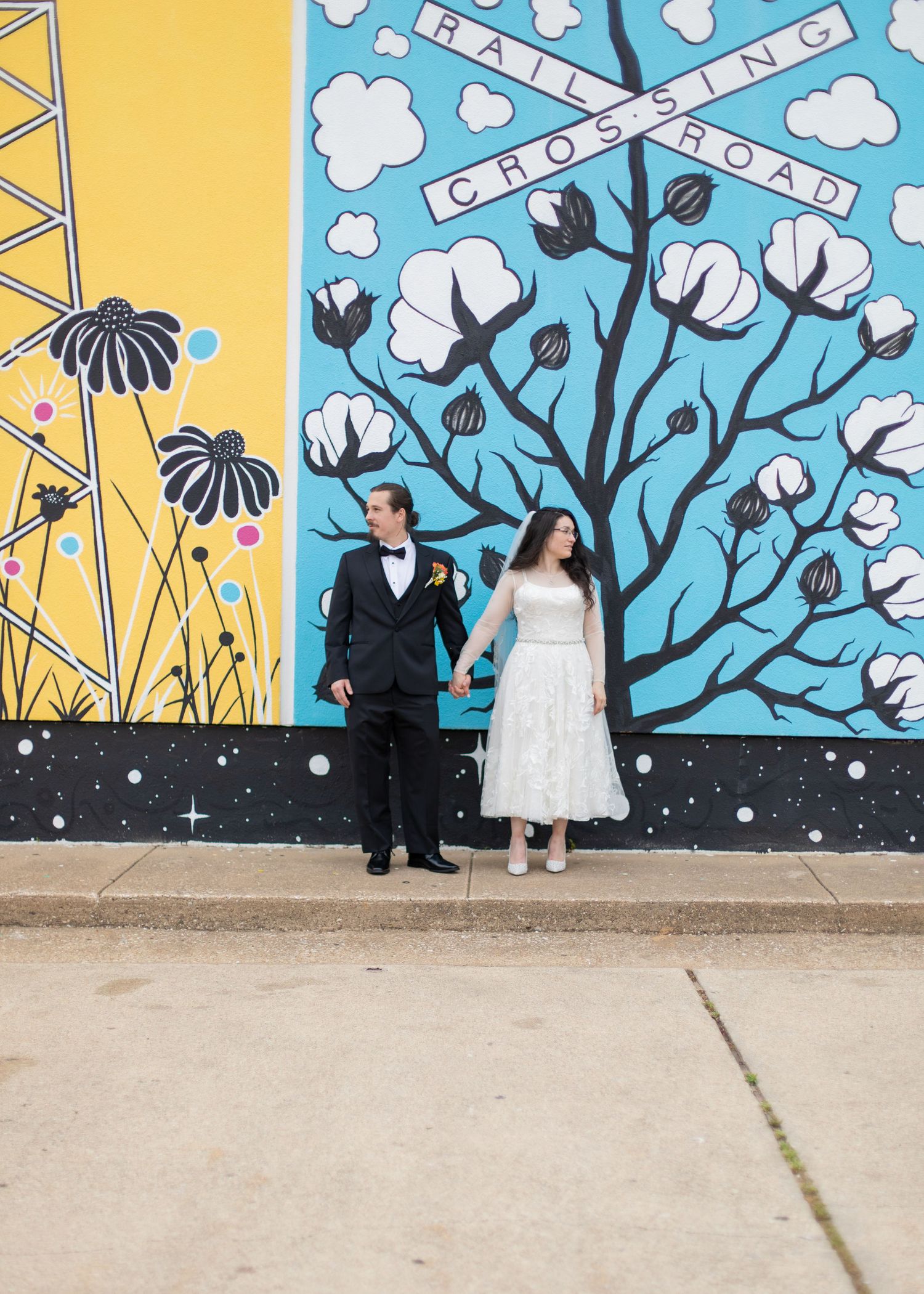 A couple stands against a vibrant mural featuring cotton plants and geometric patterns.