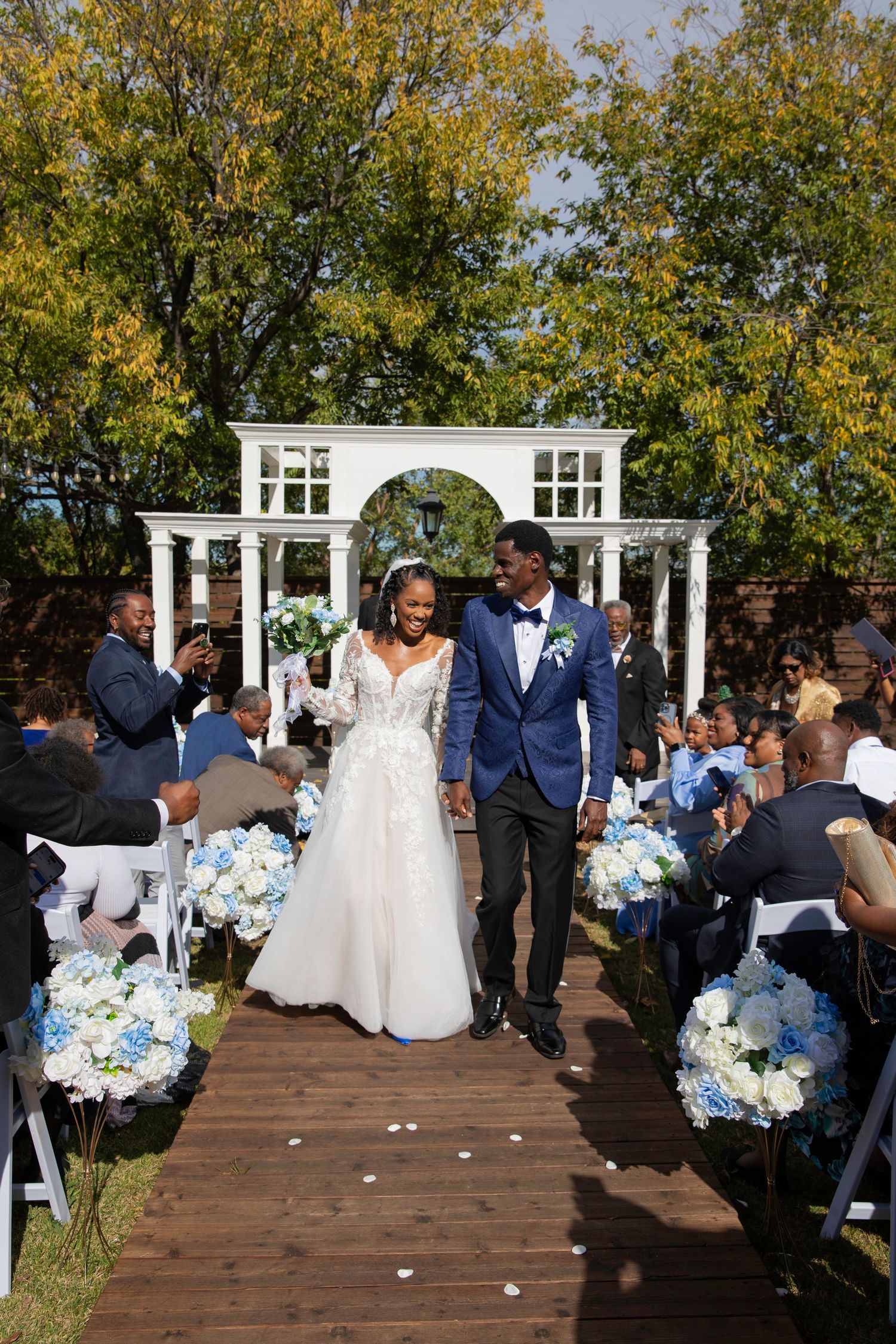 Smiling bride and groom walk back down the aisle after their outdoor wedding ceremony lined with blue hydrangea bouquets.