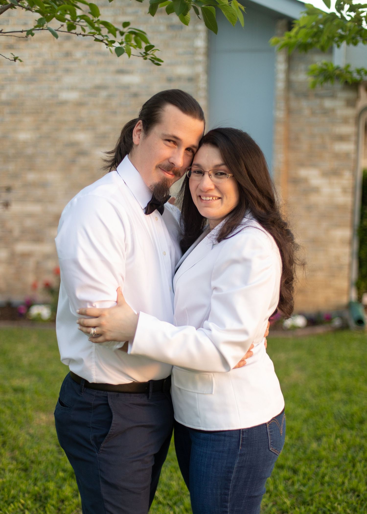 Couple in matching white tops embrace while posing in front of their home.