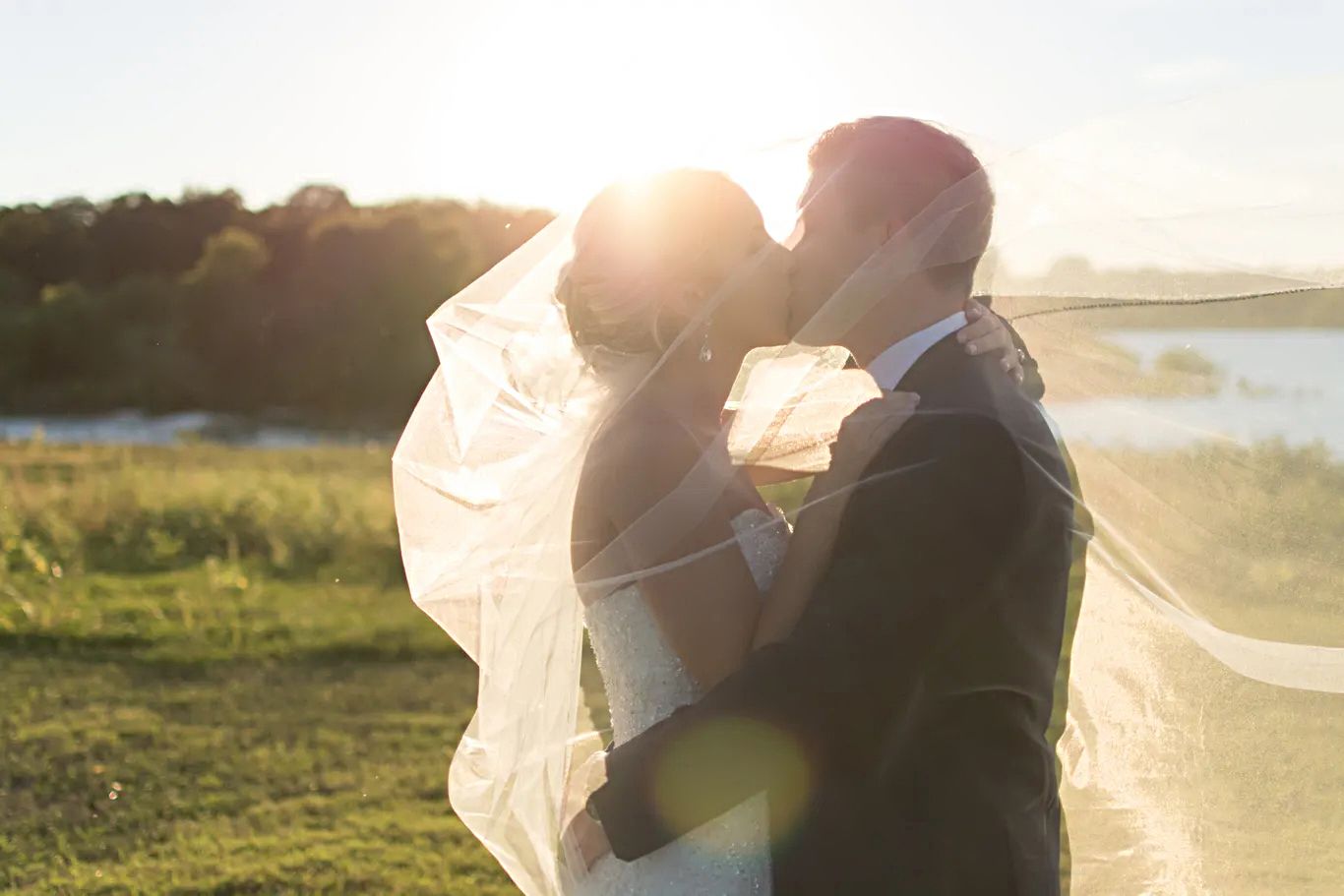 Bride and groom sharing a golden sunset kiss with wedding veil flowing in the breeze outdoors.