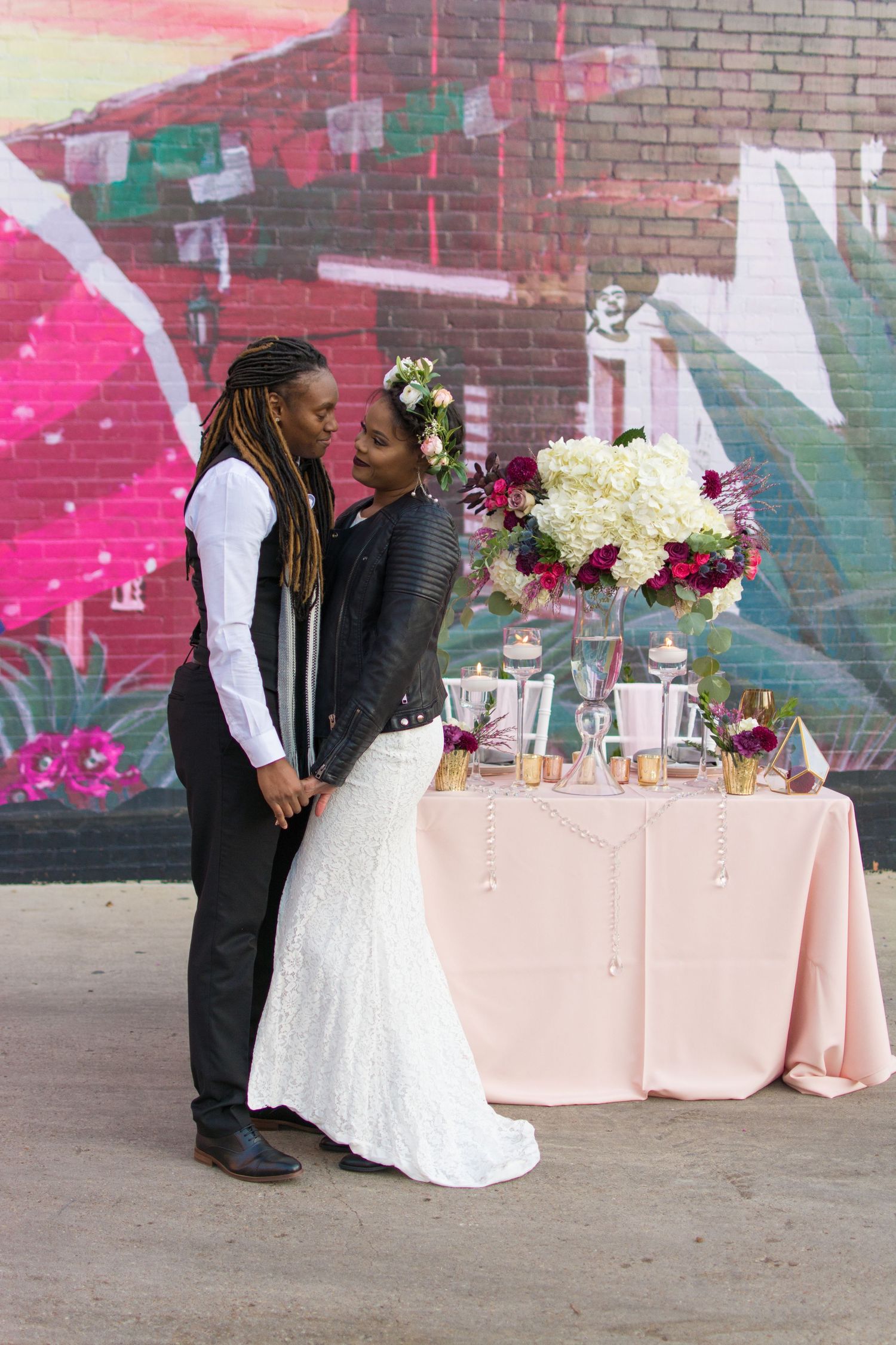 Stylish newlywed couple sharing romantic moment near floral centerpiece in front of colorful graffiti wall.