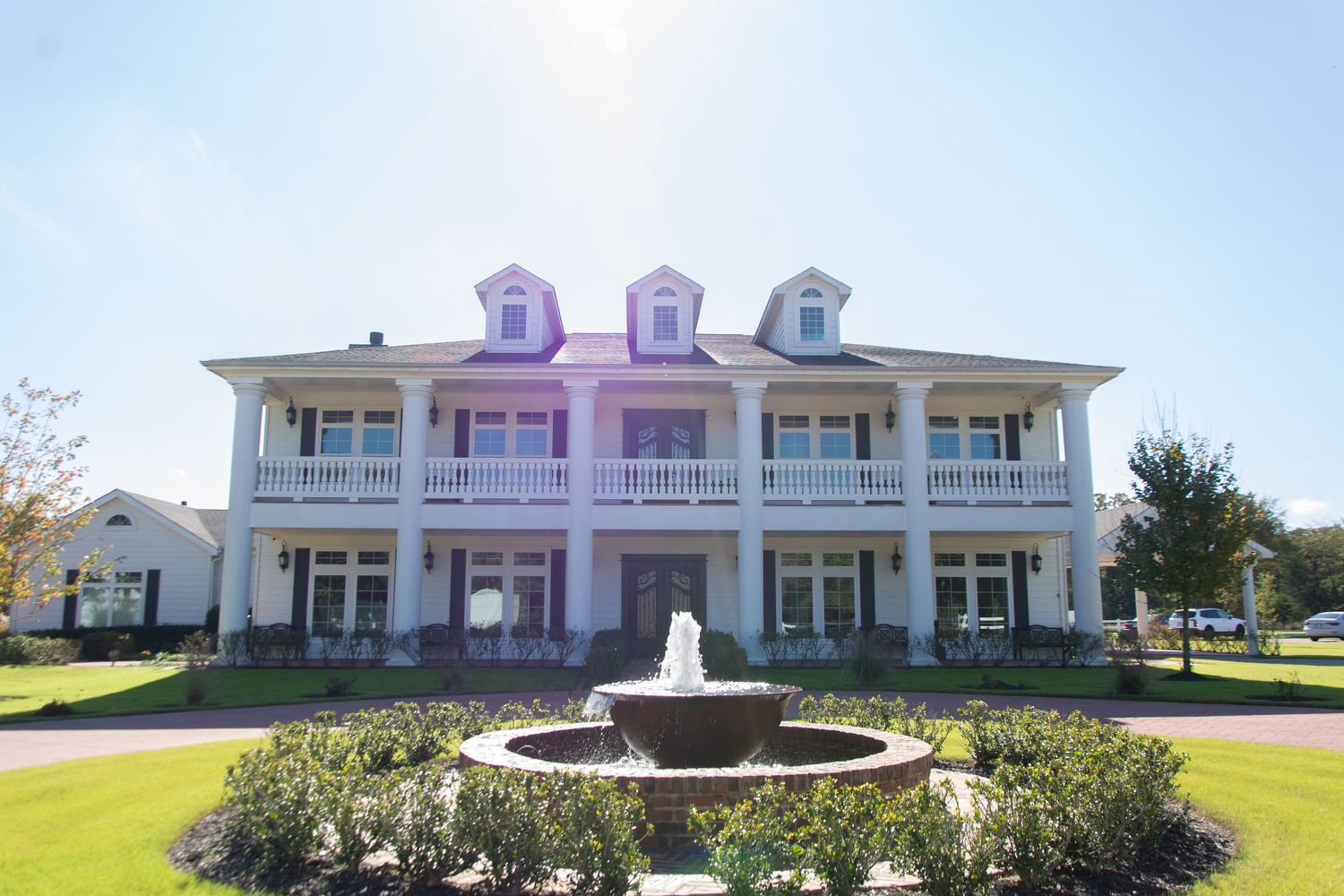 Elegant white Southern plantation-style wedding venue with fountain and manicured grounds on sunny day.