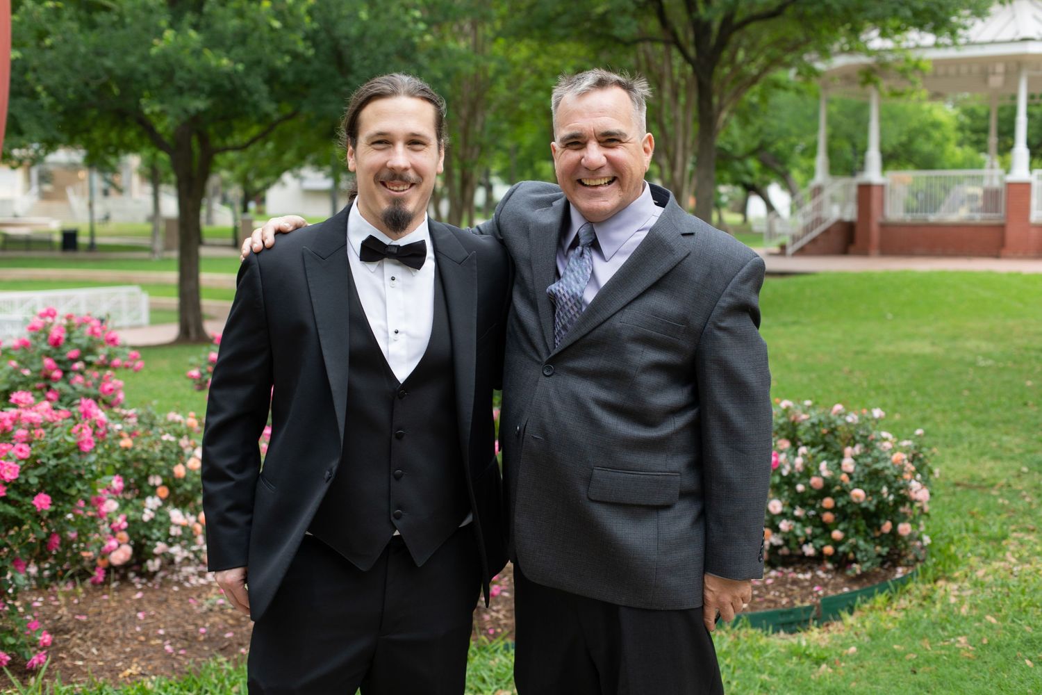 Two people in formal attire stand together smiling in a garden setting with pink flowers.