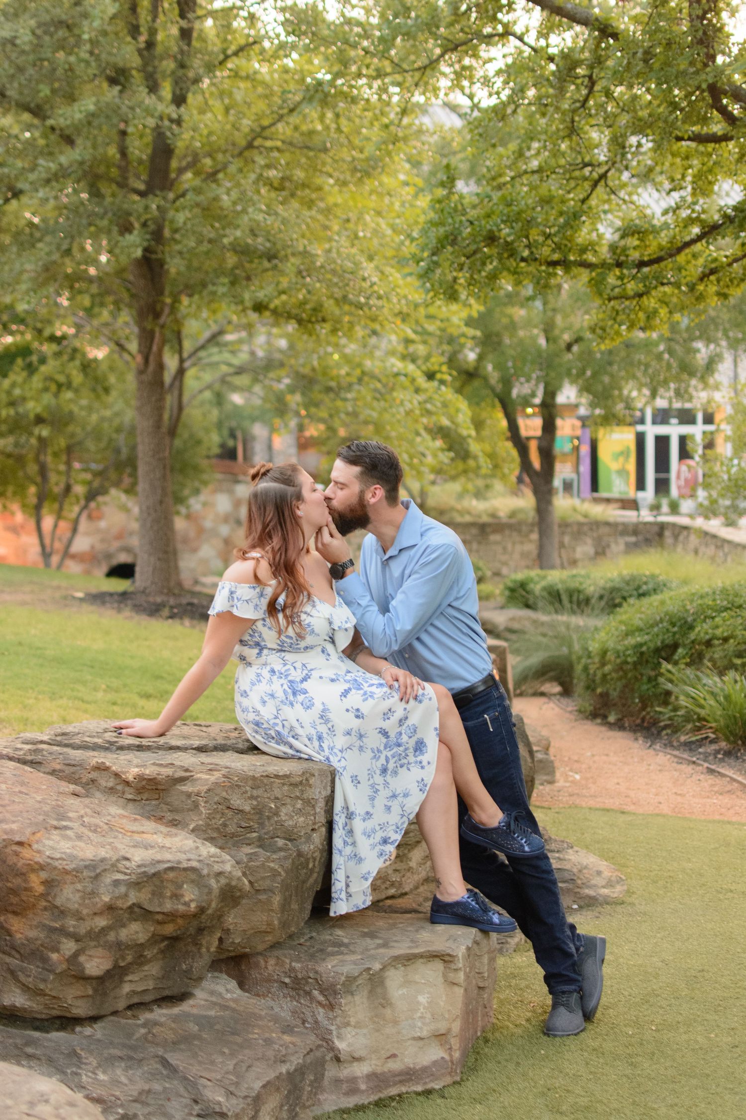 Couple sharing a gentle kiss while sitting on large rocks in a lush green park setting.
