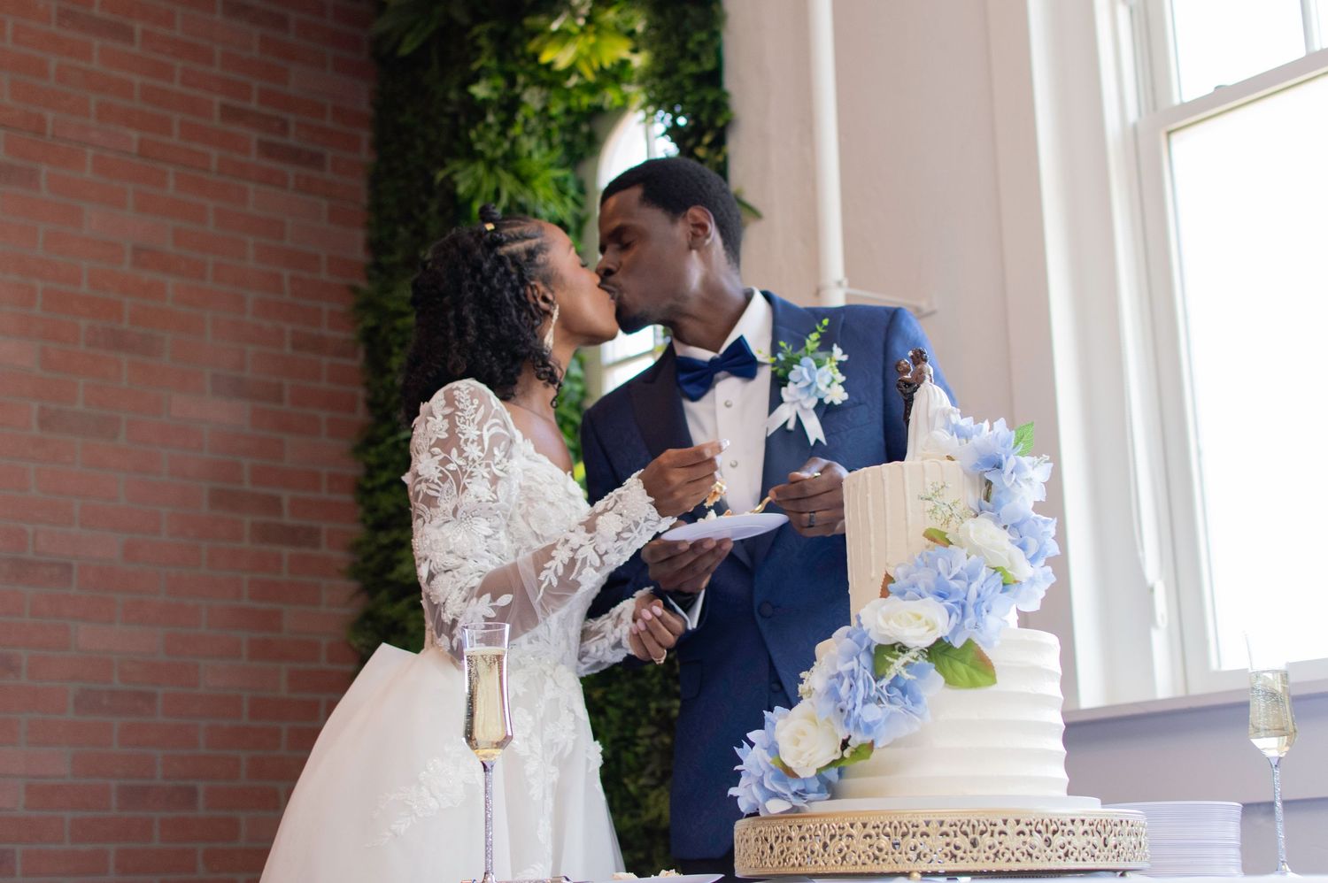 African American bride and groom share a kiss while cutting their elegant blue floral wedding cake.