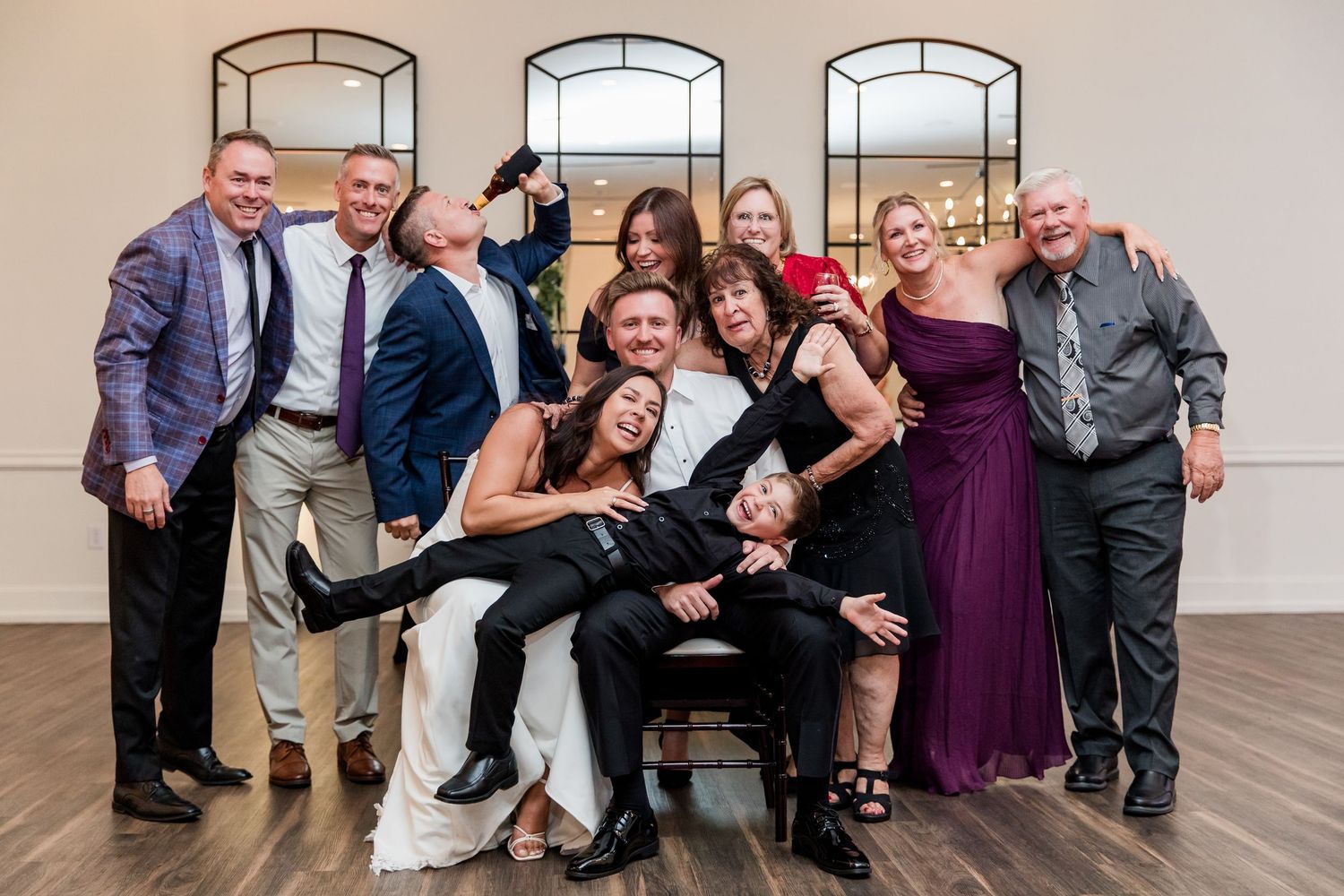 Wedding party group photo with bride and groom surrounded by smiling family members at reception.