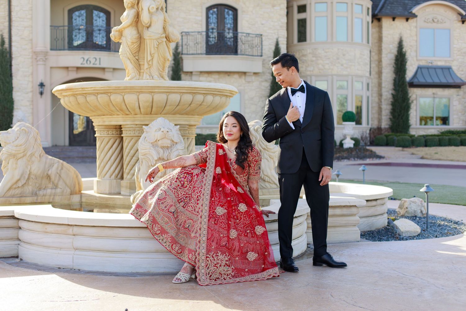 Bride in red embroidered lehenga sits by fountain as groom in black tuxedo adjusts bow tie at elegant mansion.