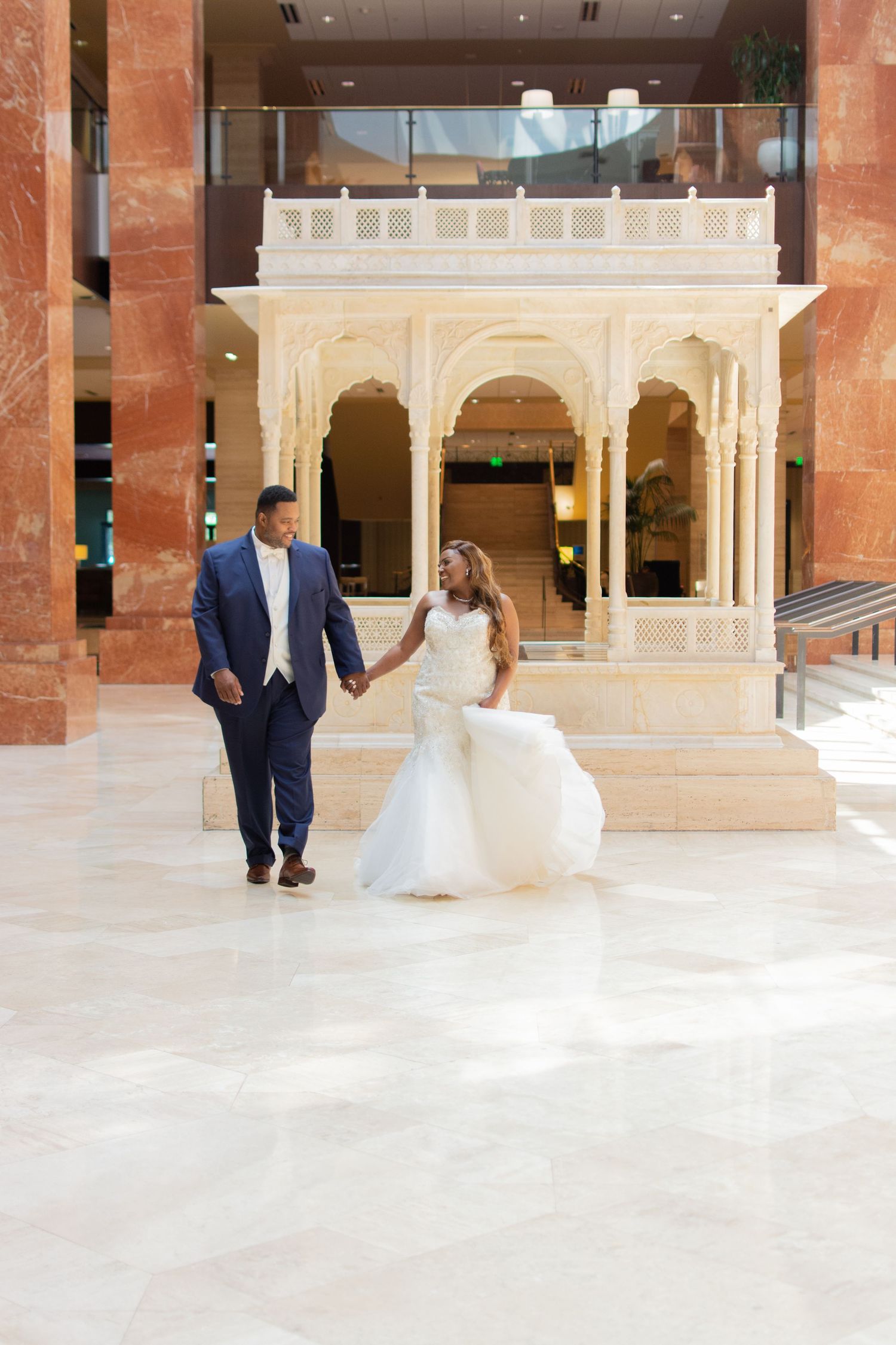 Bride in white mermaid gown and groom in blue suit walking hand-in-hand in a grand hotel lobby.