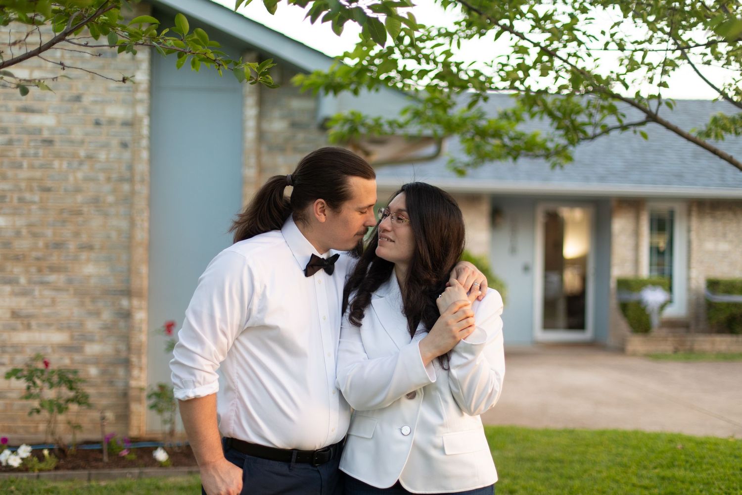 A couple in white shirts sharing an intimate moment outside a brick house with blue trim.