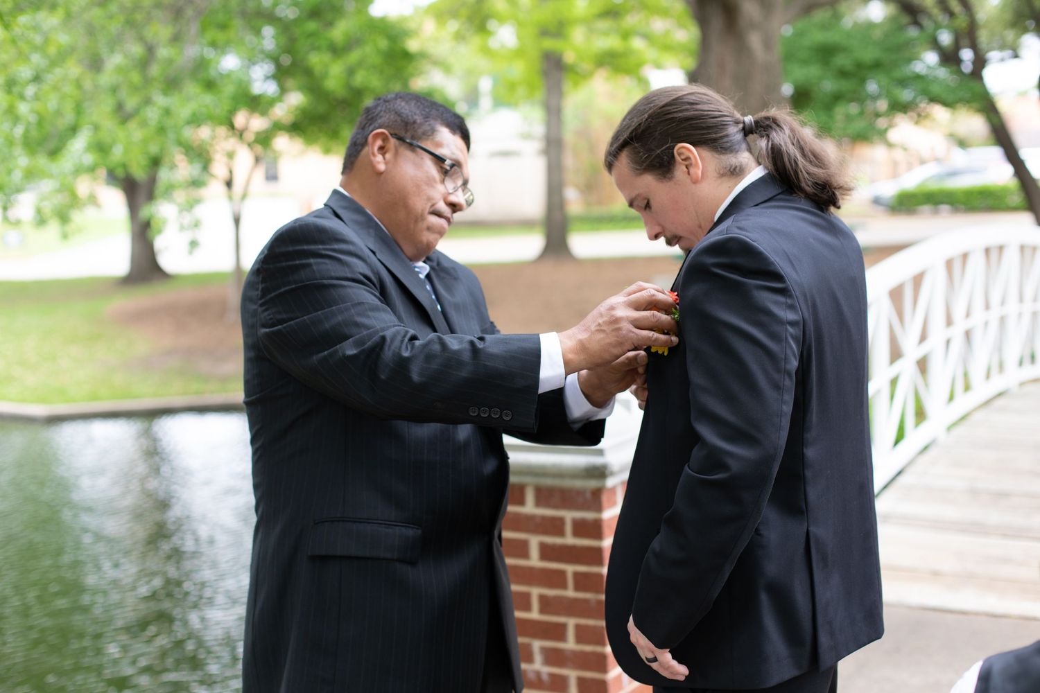 Two people in dark suits help each other with formal attire adjustments on a bridge.