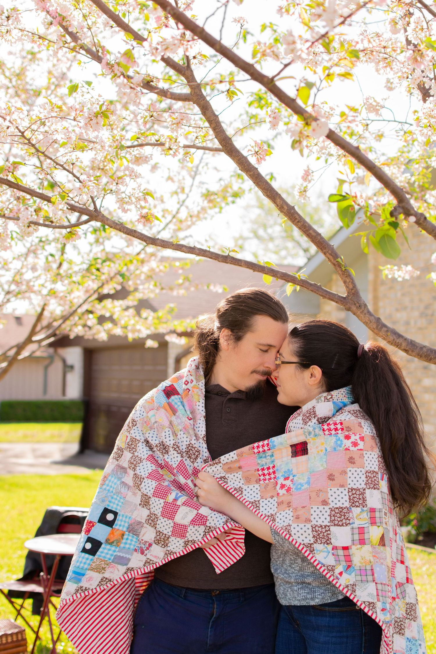 Romantic couple wrapped in colorful patchwork quilt sharing a kiss under blooming cherry blossom tree.