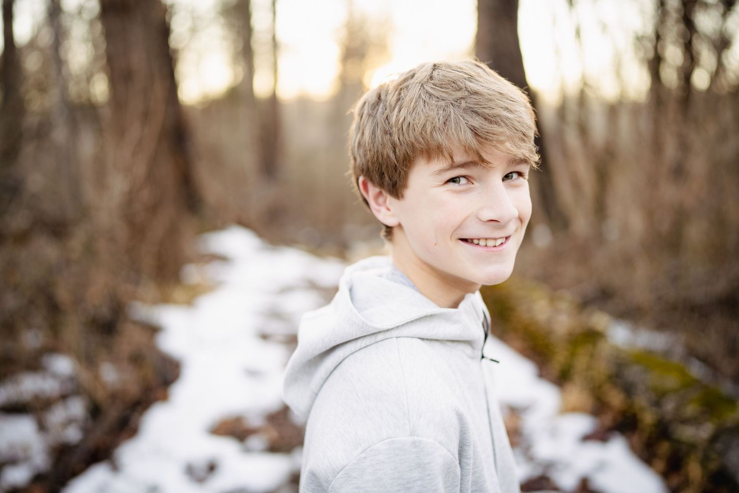 Smiling blonde teenage boy in gray hoodie poses outdoors in snowy winter forest setting.