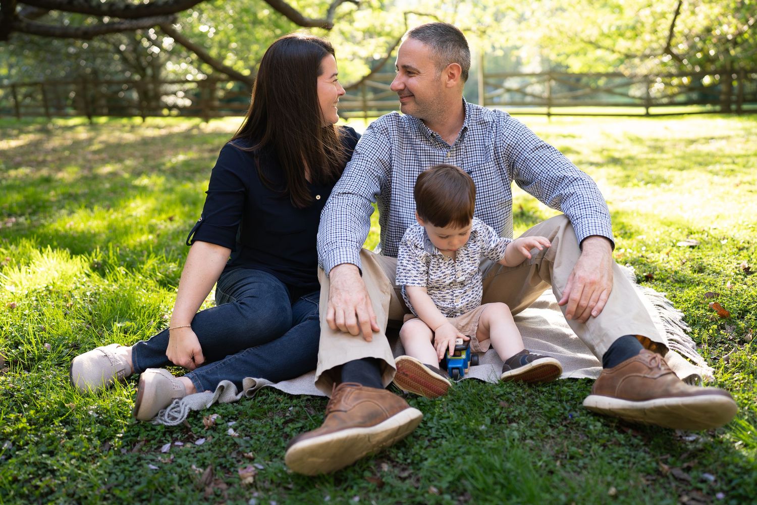 Young family of three sits together on grass in sunny park, parents smiling at each other with toddler between them.