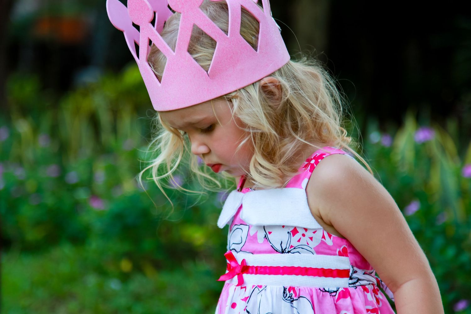 Adorable blonde girl wearing pink paper crown looks down in floral pink dress outdoors.