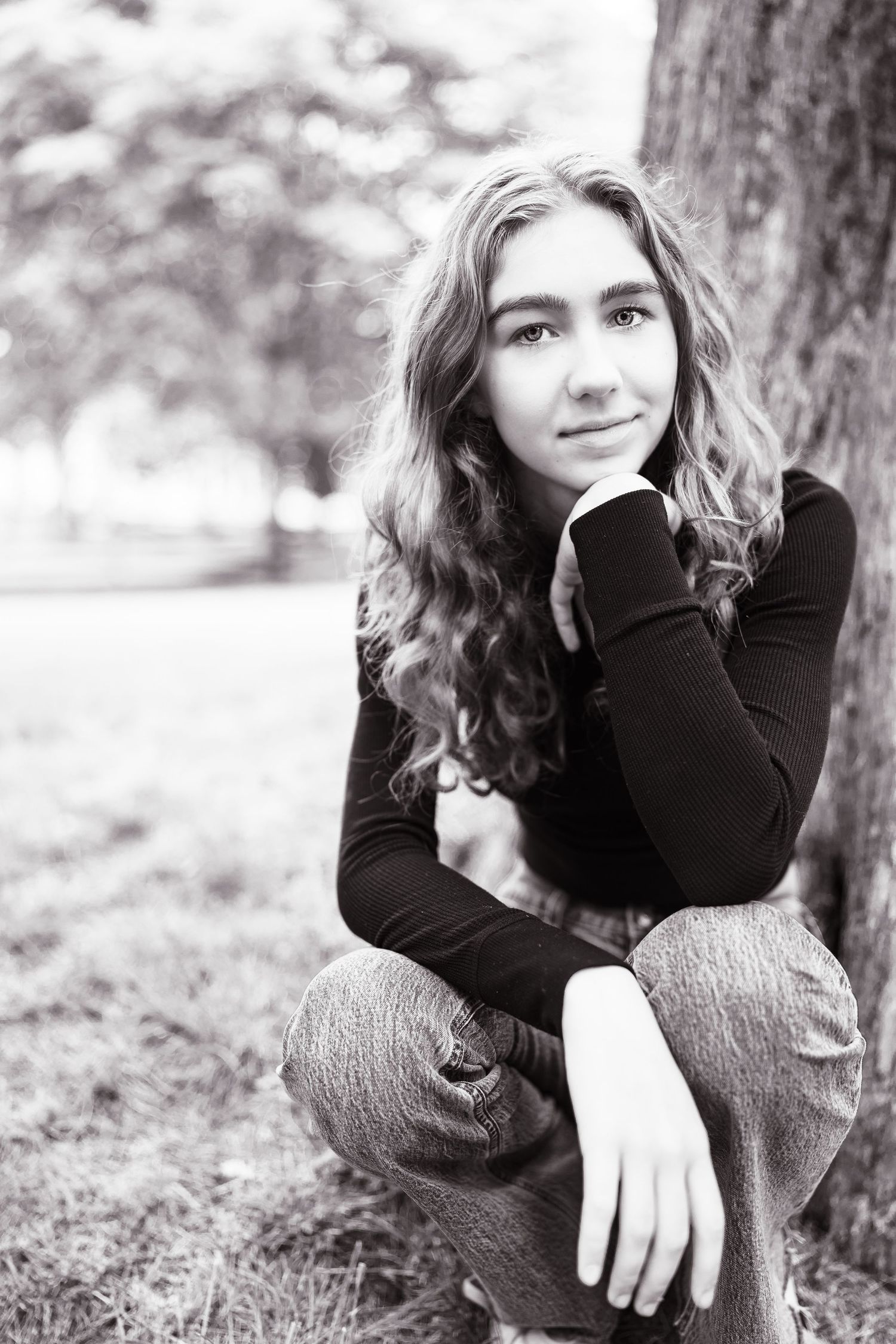 Black and white portrait of a teenage girl with curly hair sitting against a tree in a park.