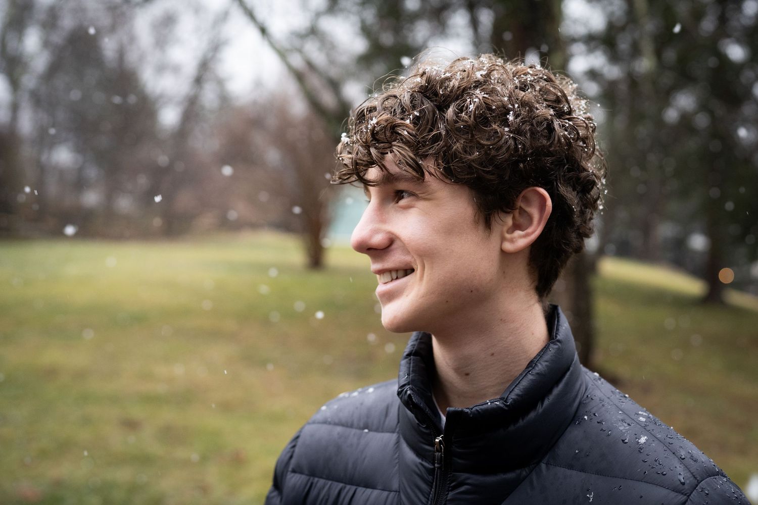 Young man with curly hair smiling outdoors in light snowfall wearing a black puffer vest.