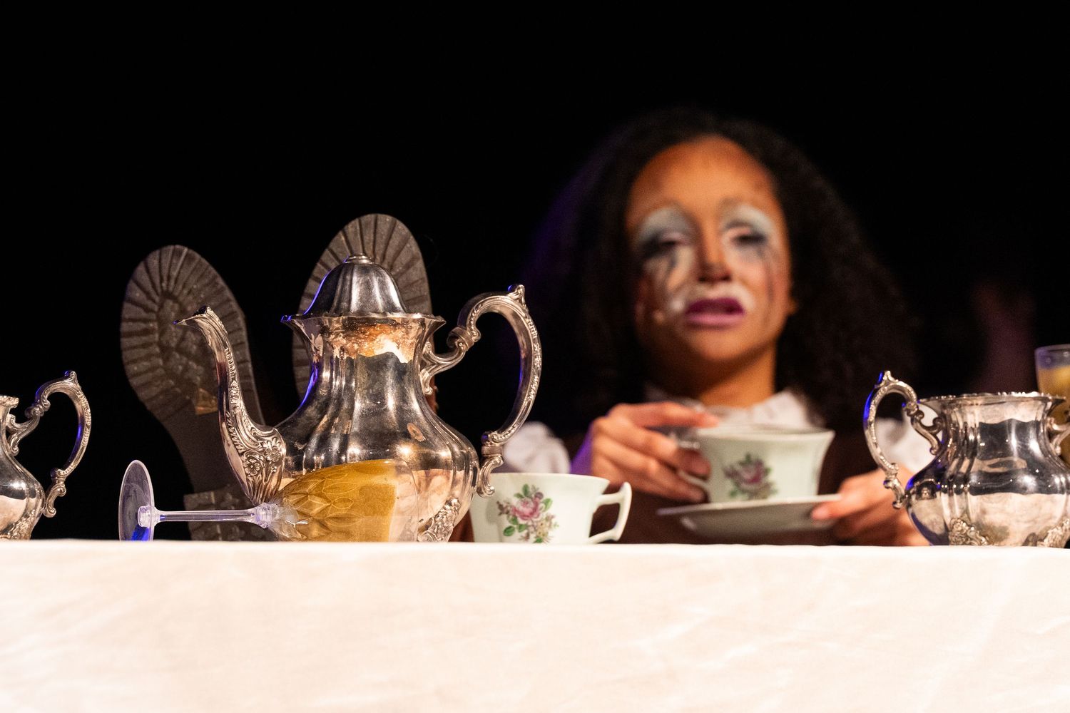 Actress in elaborate face makeup peers over a silver tea service during a stage performance.