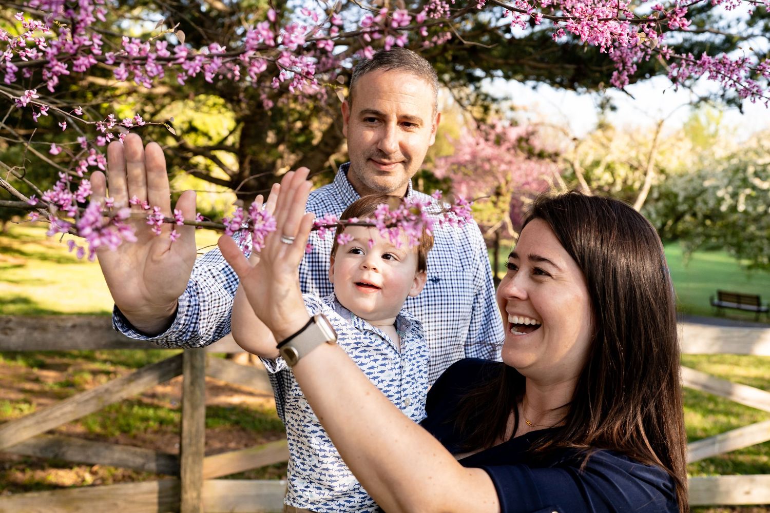 Family of three enjoying spring blossoms outdoors, mother holding baby while father smiles behind flowering pink tree.