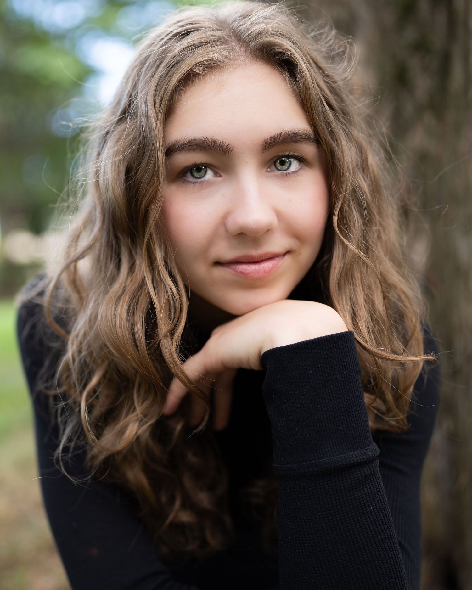 Young woman with wavy blonde hair and green eyes posing outdoors near a tree in a black turtleneck.