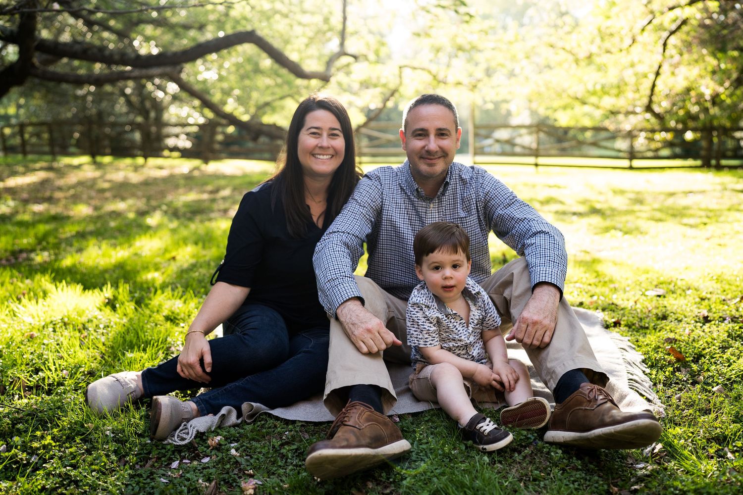 Family of three smiling and sitting on green grass in a sunlit park.