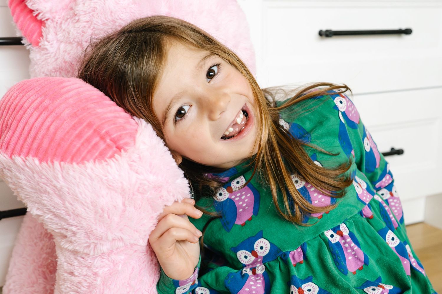 Smiling young girl in green owl-print dress hugging a large pink stuffed animal.