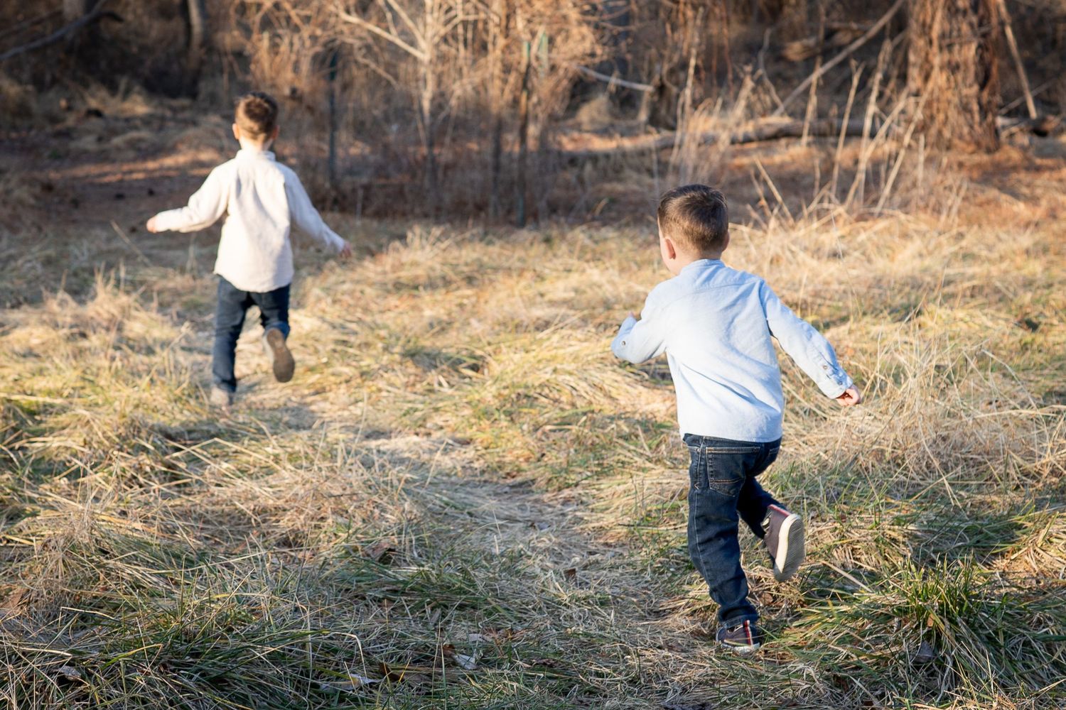 Two young boys in white shirts run freely through an open field with dry grass and bare trees.