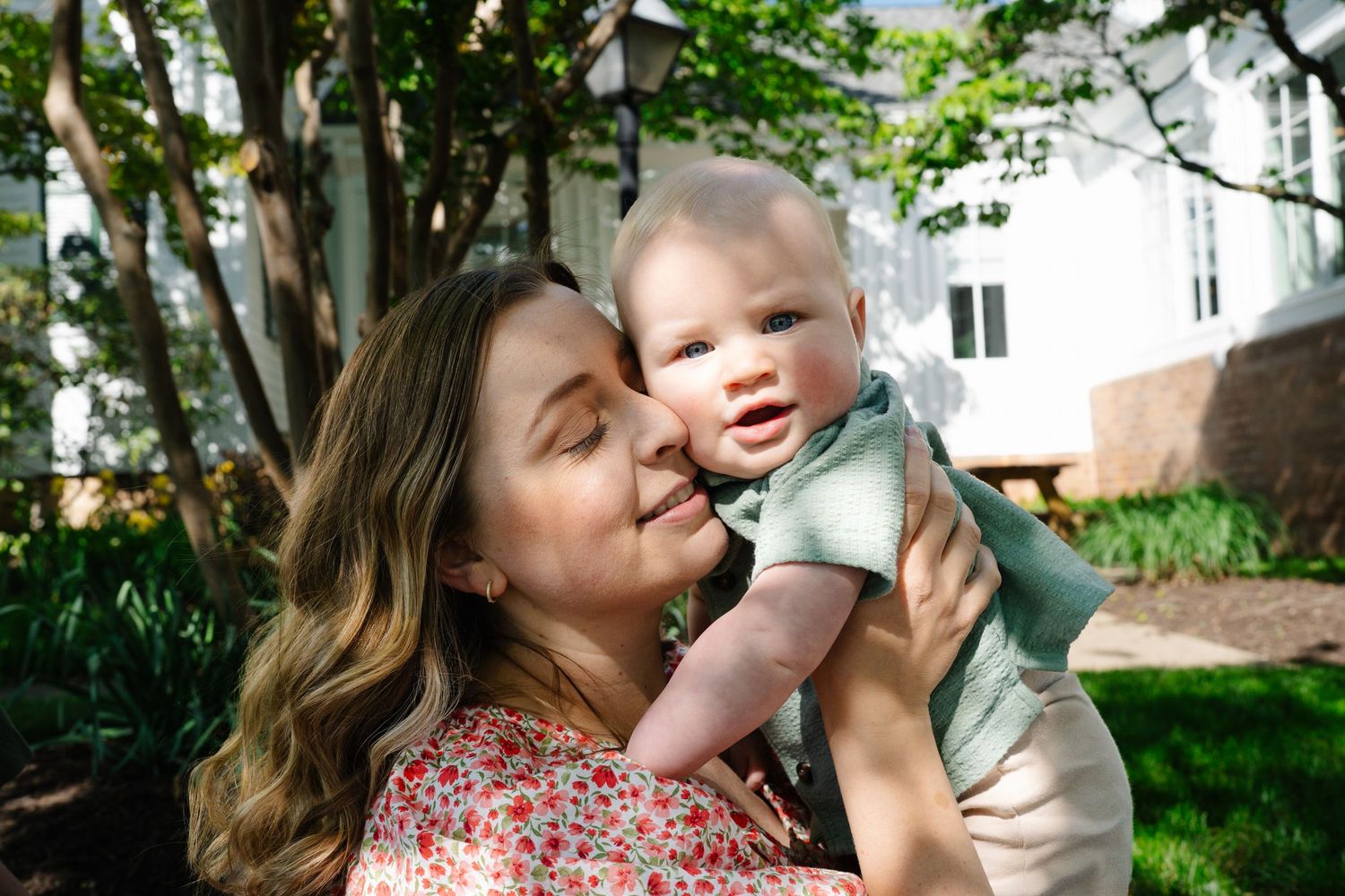 Joyful mother holding and kissing her smiling baby outdoors near a white building with greenery.