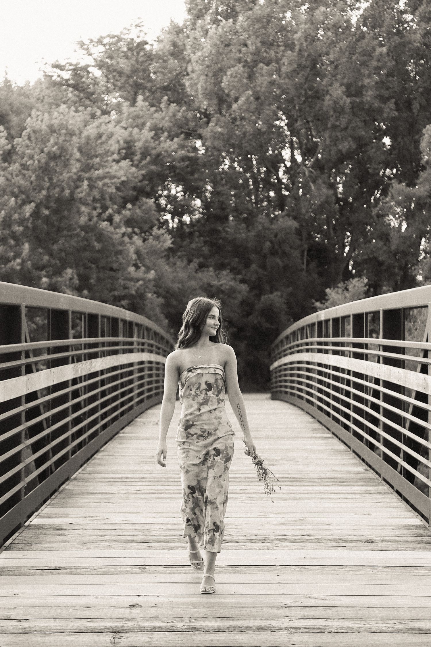 A person in a floral dress walks along a wooden bridge pathway surrounded by trees.