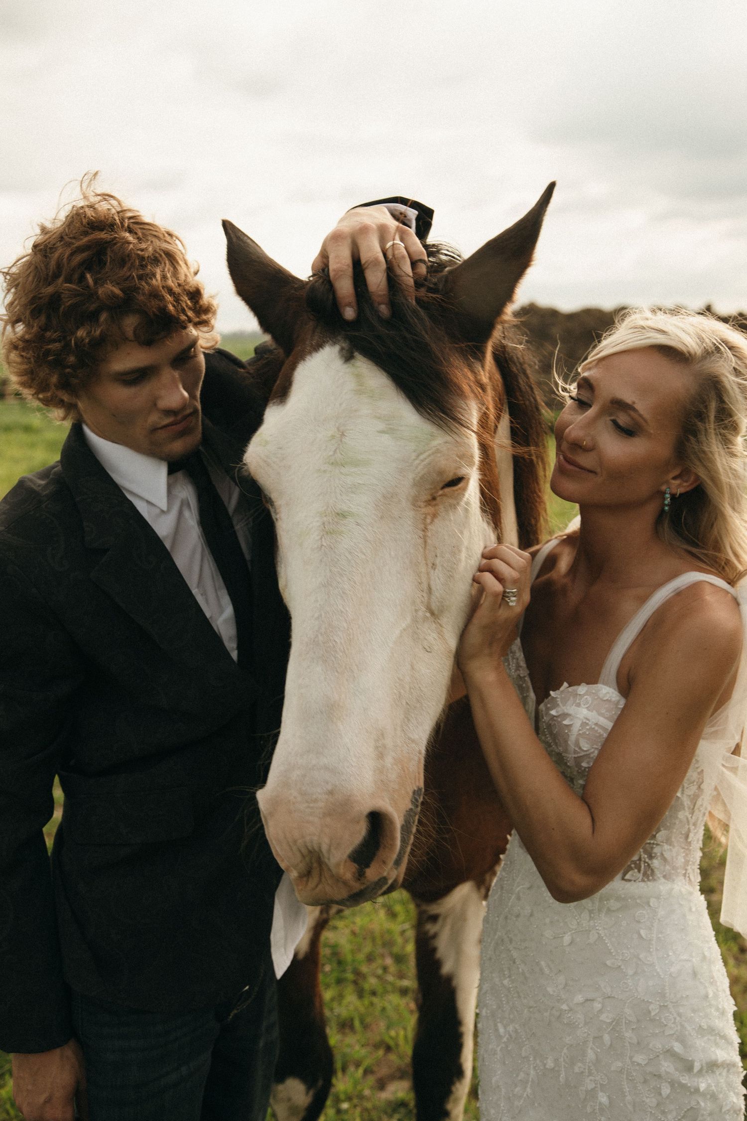 Bride and groom gently petting a pinto horse together outdoors on wedding day.