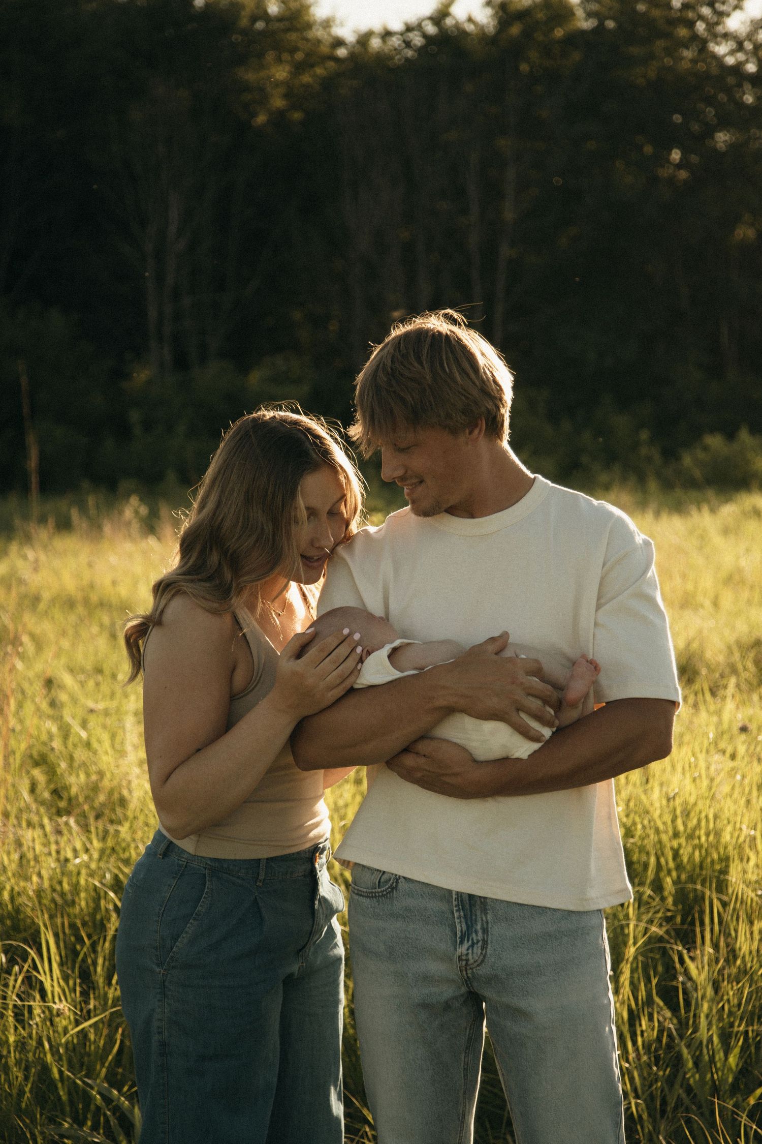 Happy couple holding newborn baby in sunlit green field during golden hour photography session.