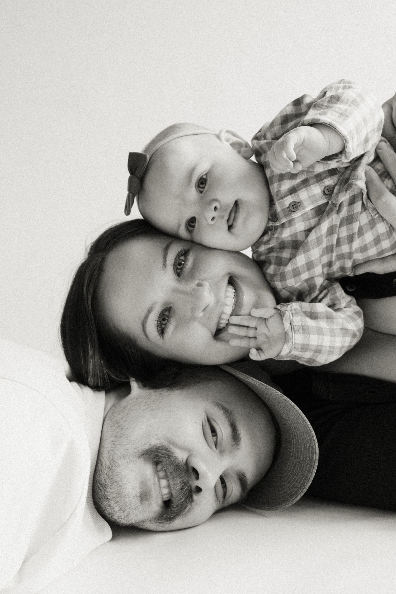 Black and white family portrait of smiling parents lying down with happy baby resting on top, studio photo.