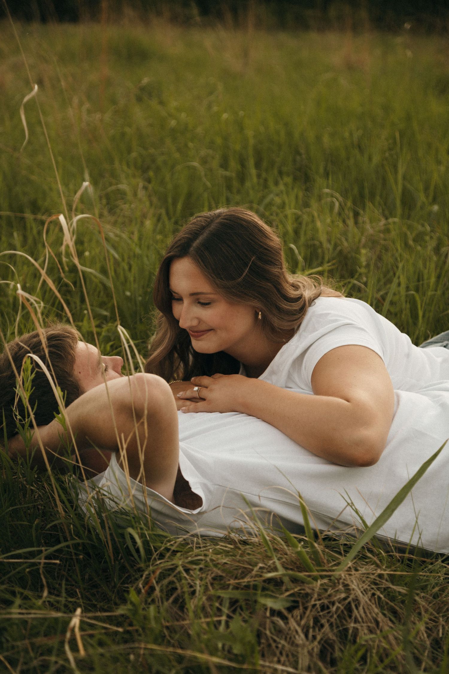 Romantic couple lying together in tall green grass, gazing lovingly at each other outdoors.