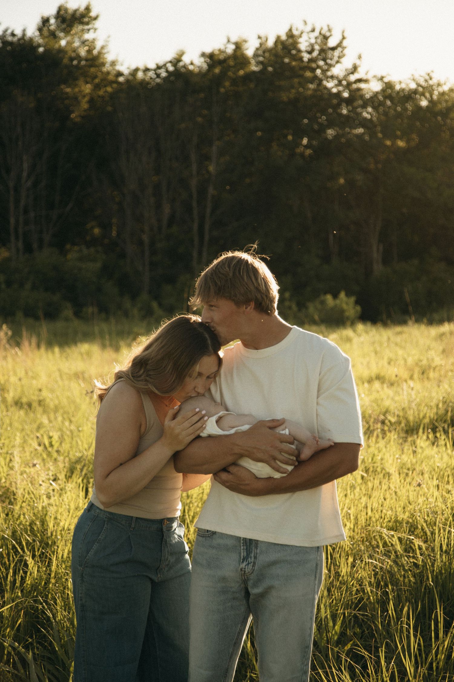 Young couple standing in golden hour field, tenderly cradling their newborn baby together.