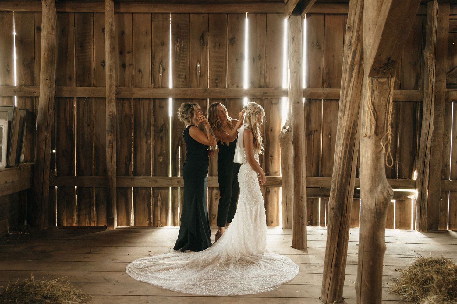 Bride in lace gown being helped by bridesmaids in dark dresses inside a rustic wooden barn wedding venue.
