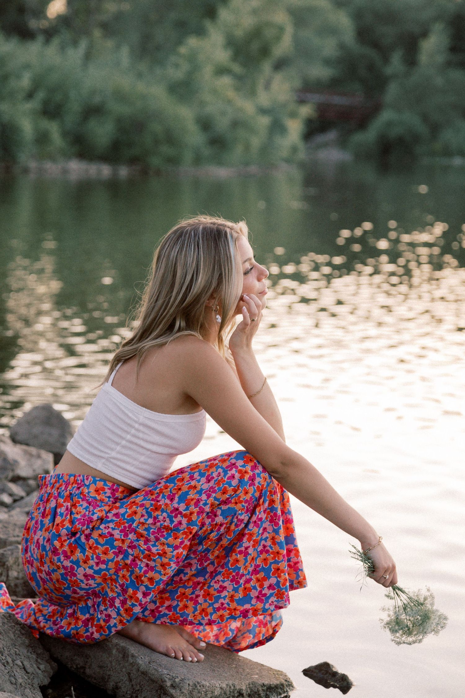 Figure in red floral skirt sits on rocks by a tranquil lake during golden hour.